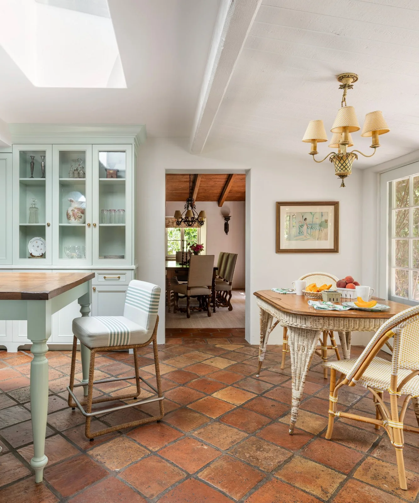 A terracotta grid continues beneath striped kitchen stools and a woven breakfast table beside pale cabinets.