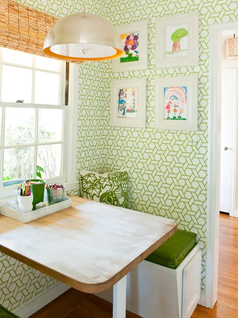 Green-cushioned fitted bench against green-and-white geometric wallpaper, with a rounded wooden tabletop, white bench base, framed children’s drawings, a woven window shade, and domed pendant.