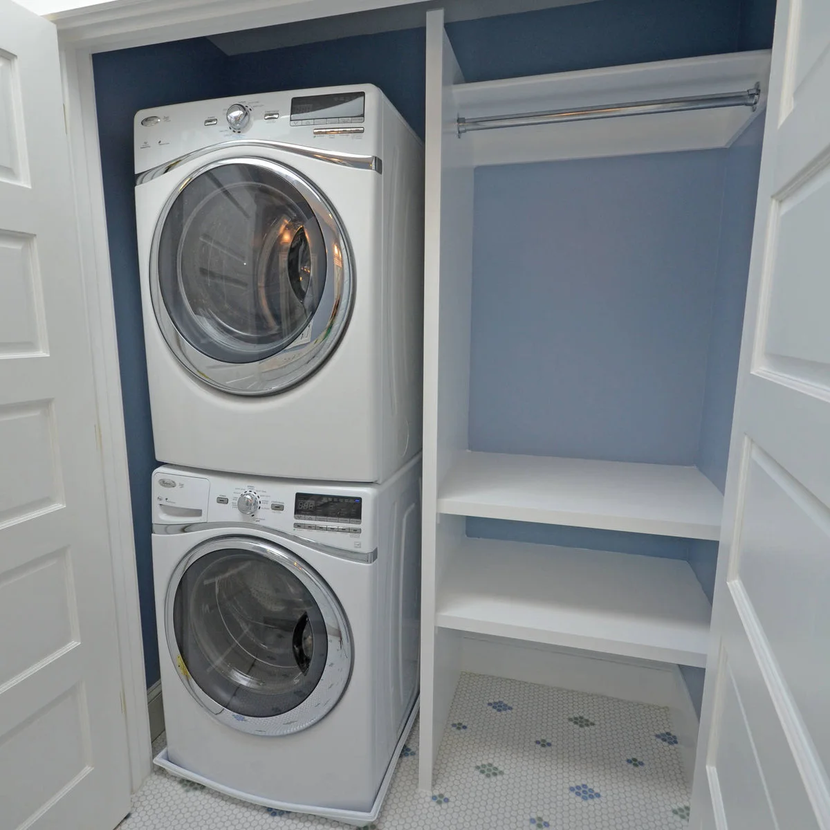 Compact laundry closet with stacked appliances, blue walls, two open white shelves and a hanging rail to the right