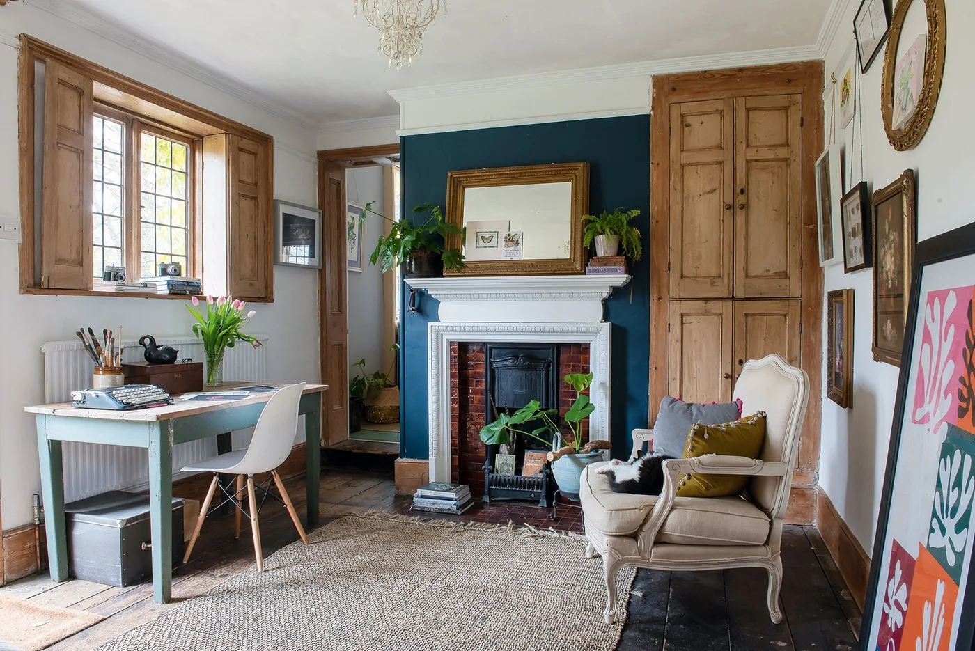 A white fireplace with a dark insert and red side tiles stands against a teal chimney breast in a study with timber shutters.