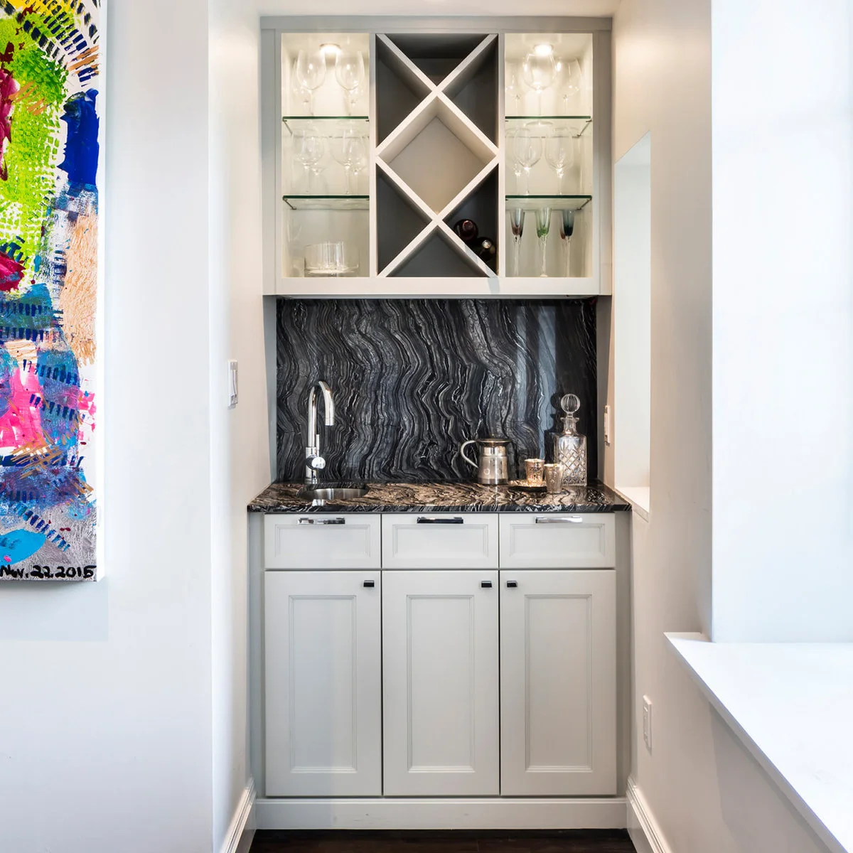 Narrow white mini bar with dark swirled backsplash, small sink and upper display shelves around diagonal bottle storage