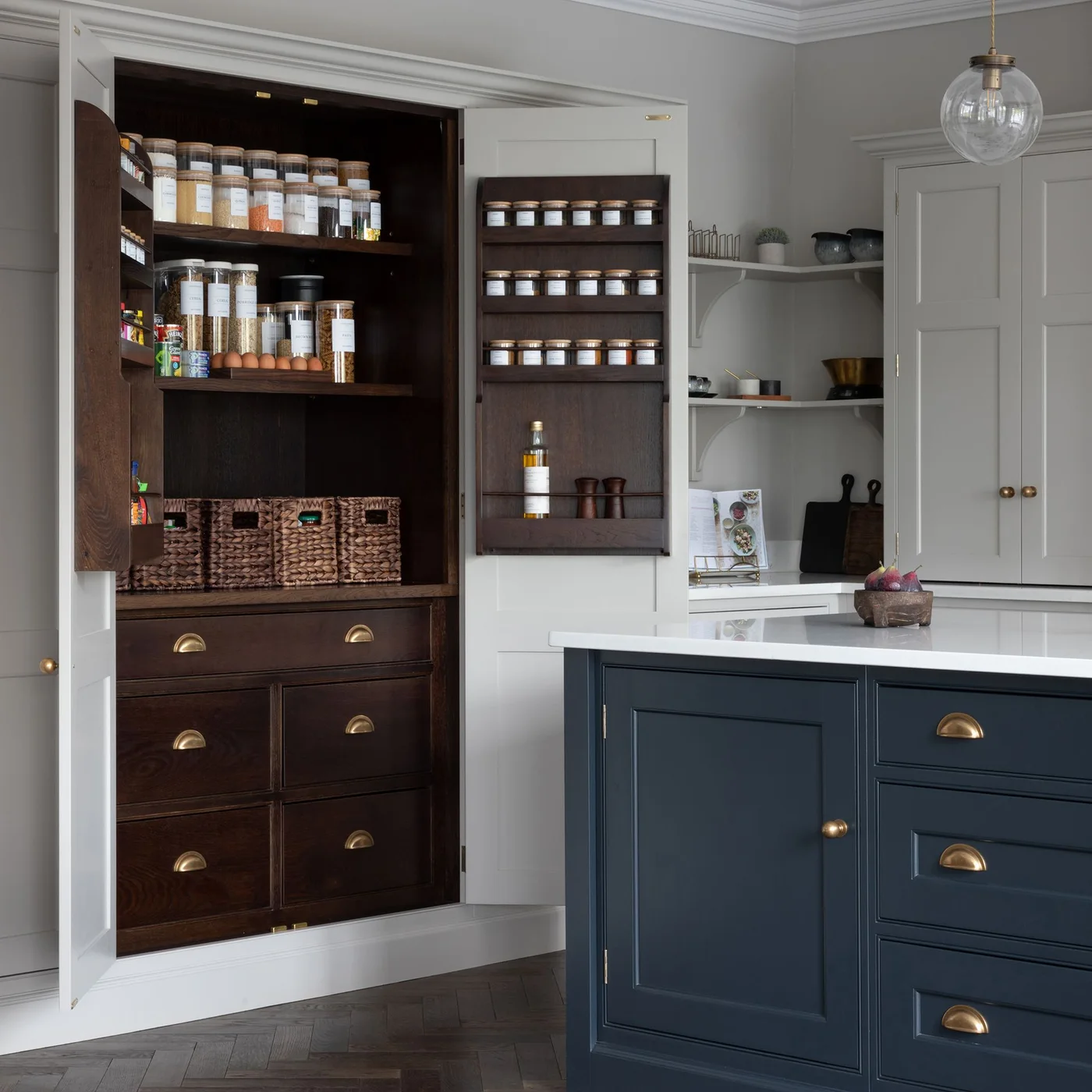 A dark blue-green island stands beside an open pantry with wood-toned shelves, jars, baskets, door-mounted spice racks and lower drawers