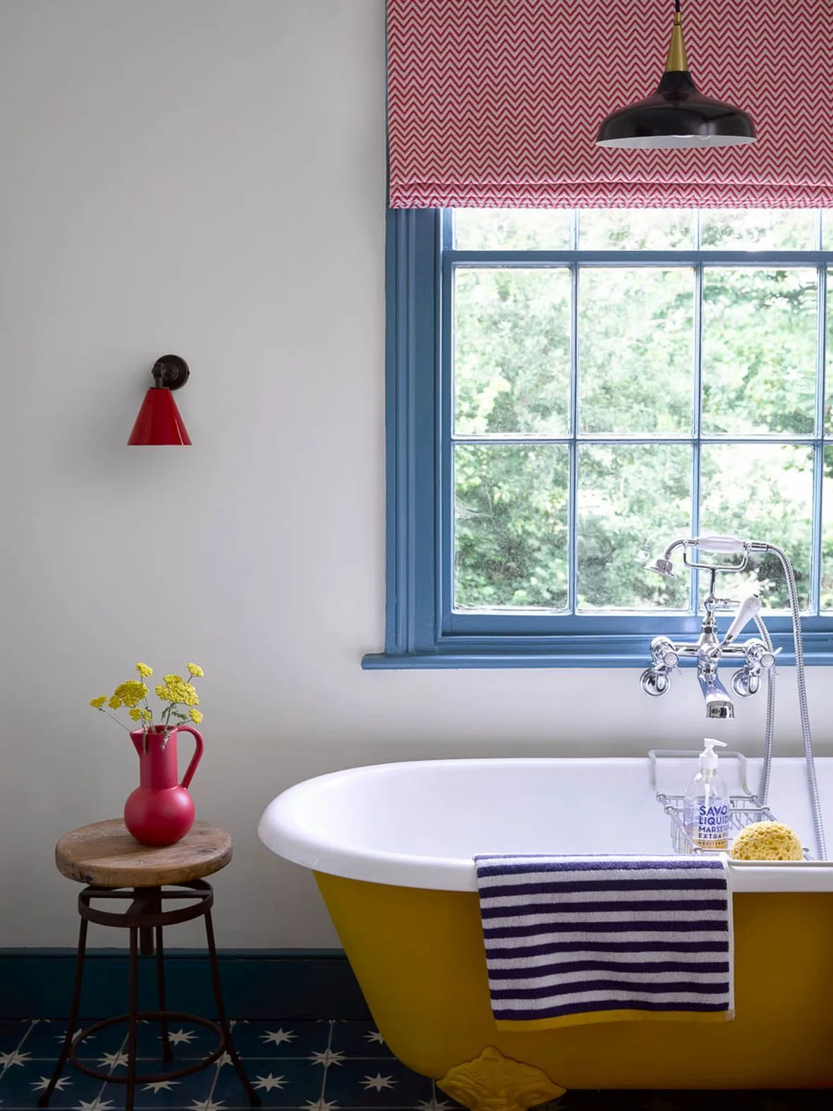 Bathroom with a yellow claw-foot bath, blue window frame, red patterned blind, striped towel and dark star-patterned floor.
