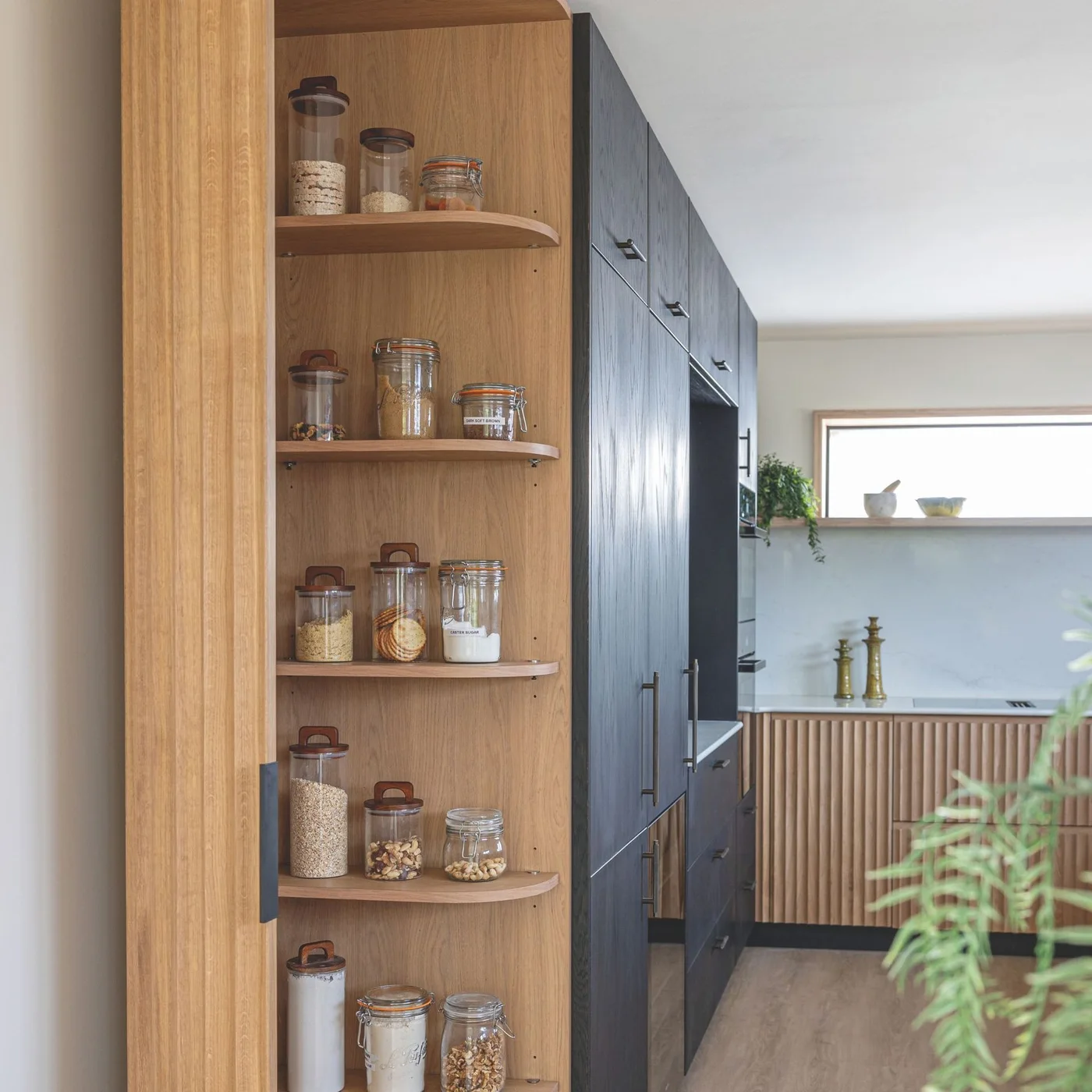 An open curved oak end cupboard holds labeled glass jars beside black tall kitchen cabinets and a fluted lower run.