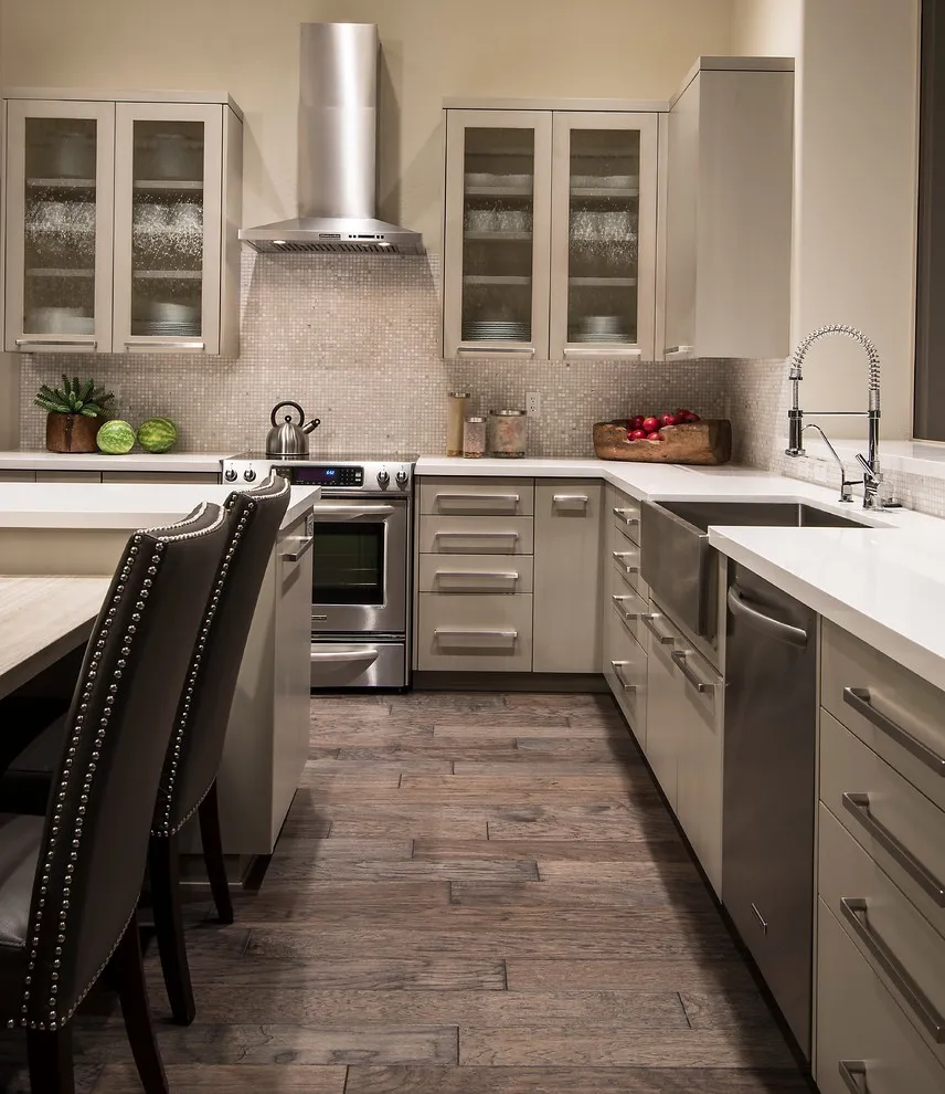Beige paneled cabinets extend around a long kitchen with glass uppers and dark dining chairs