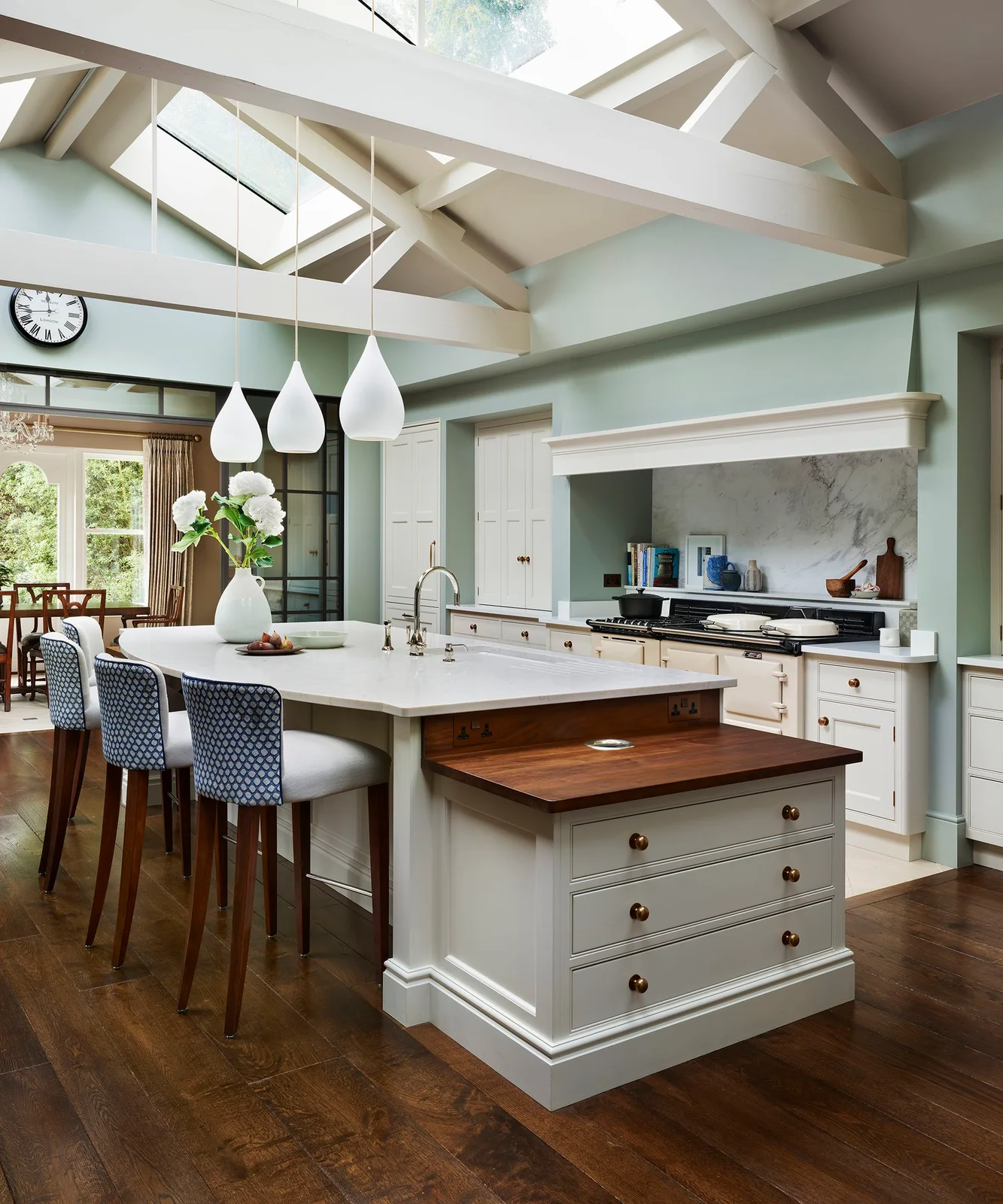 Warm pale framed cabinets and a cream range surround an island with a lower timber-topped drawer section under roof glazing.