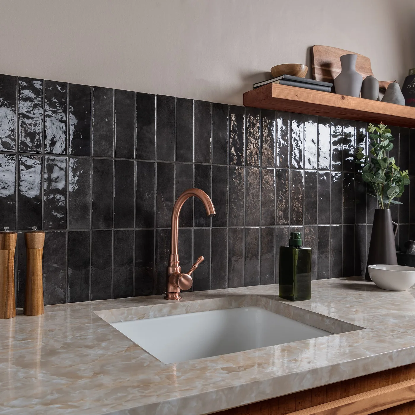 Glossy black tiles are stacked vertically behind a pale kitchen counter and copper-toned tap beneath a wooden shelf