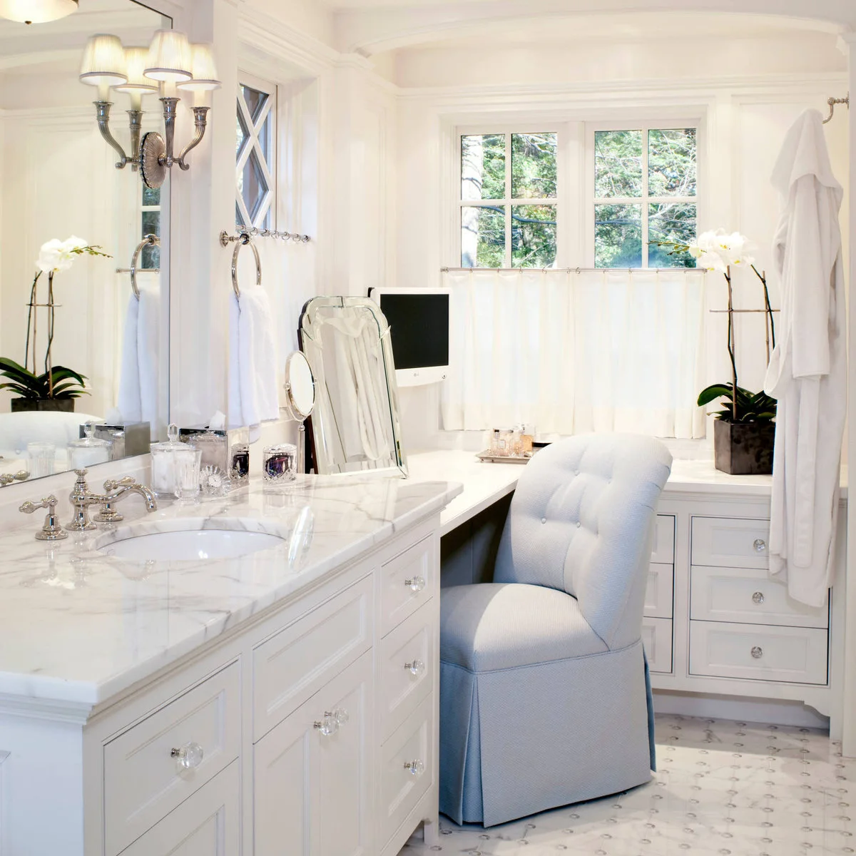 Short white curtains cover the lower window behind a bathroom dressing counter with a pale blue upholstered chair.