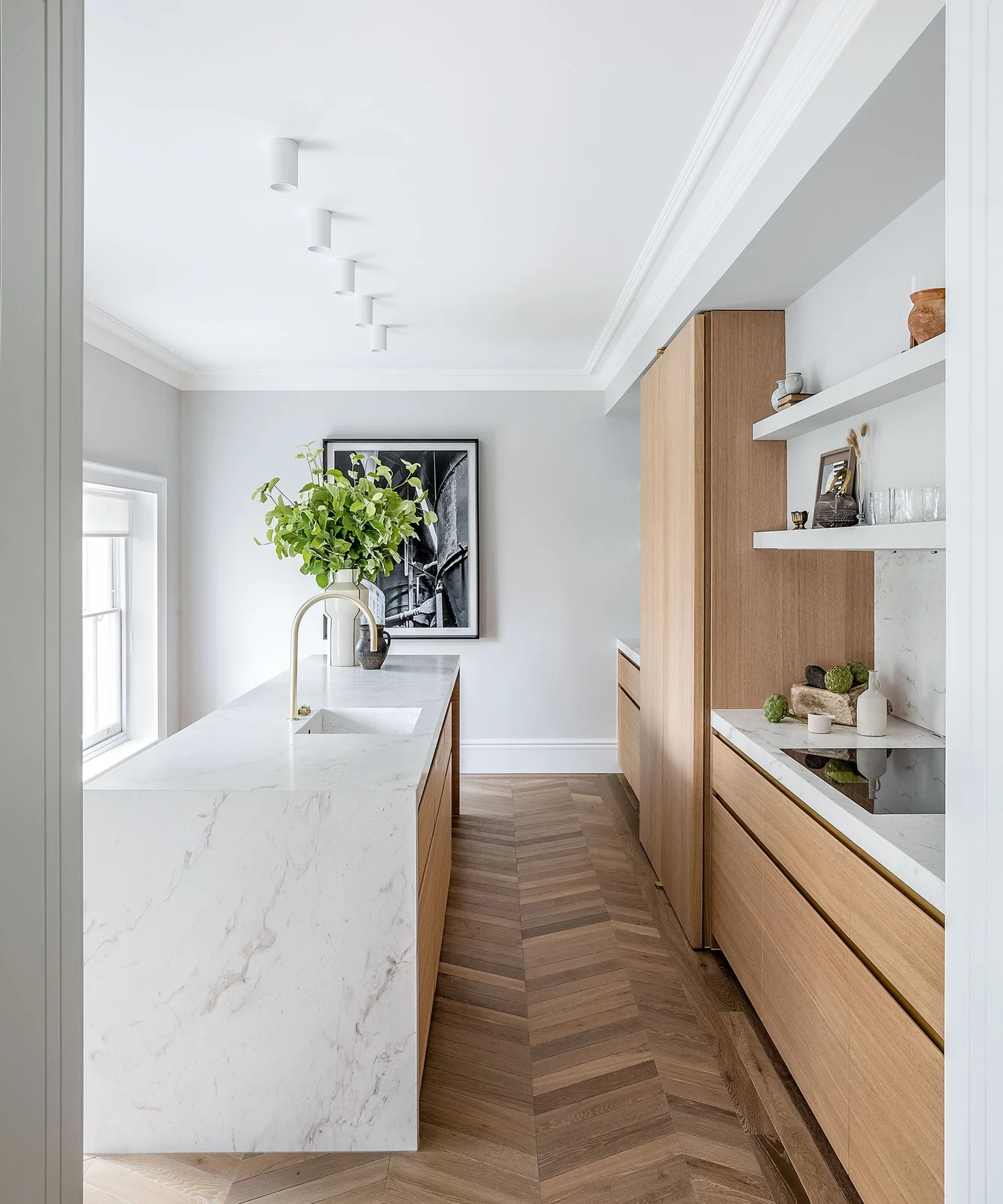 Light oak slab cabinets and drawers face a pale waterfall counter in a narrow white kitchen with a herringbone floor.