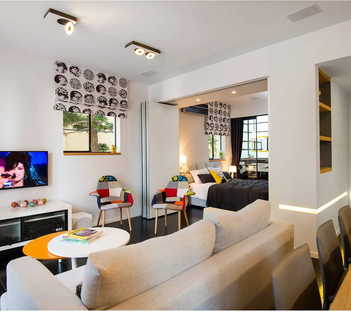 A small apartment has a pale sofa facing a wall television, colorful chairs near a bedroom opening and dining chairs at the edge behind the sofa.