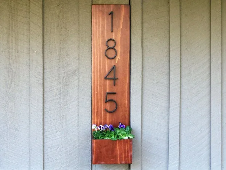 Vertical wood house-number board with small flower planter against gray board siding