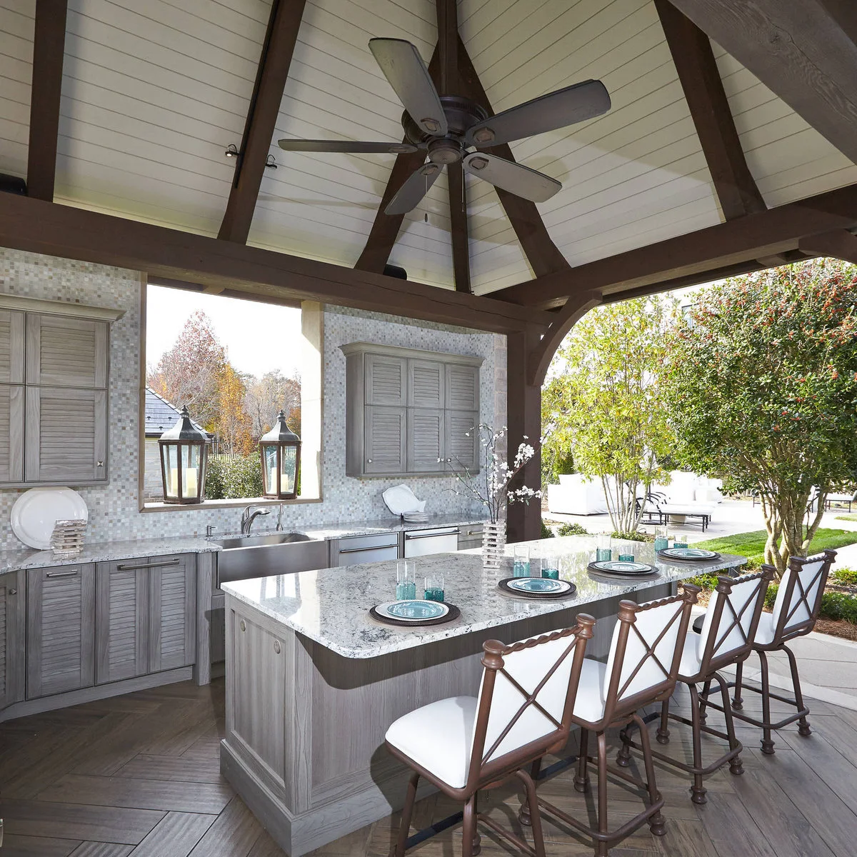 Outdoor preparation kitchen with rear sink, gray cabinets, pale mosaic wall and island seating with cross-back stools