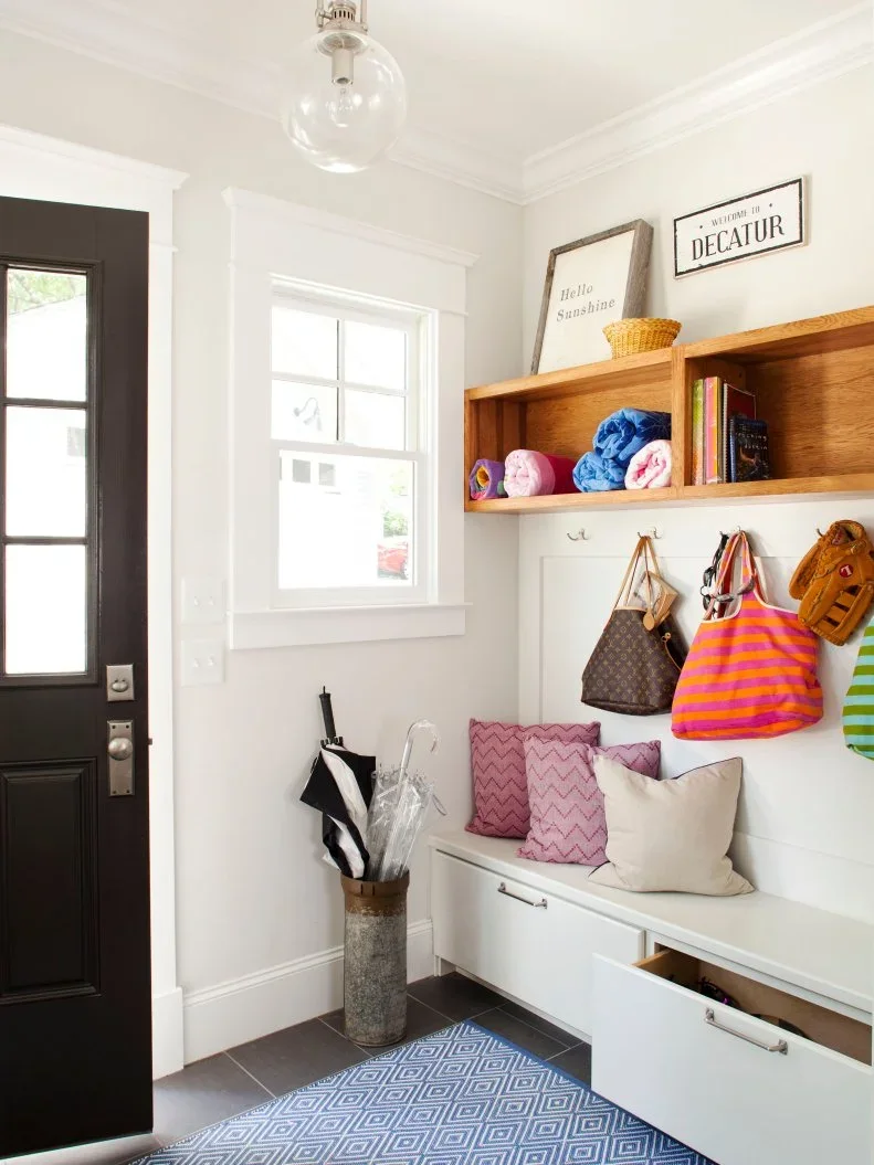 A white bench with two deep drawers sits beneath wood cubbies and coat hooks beside a black glass door.