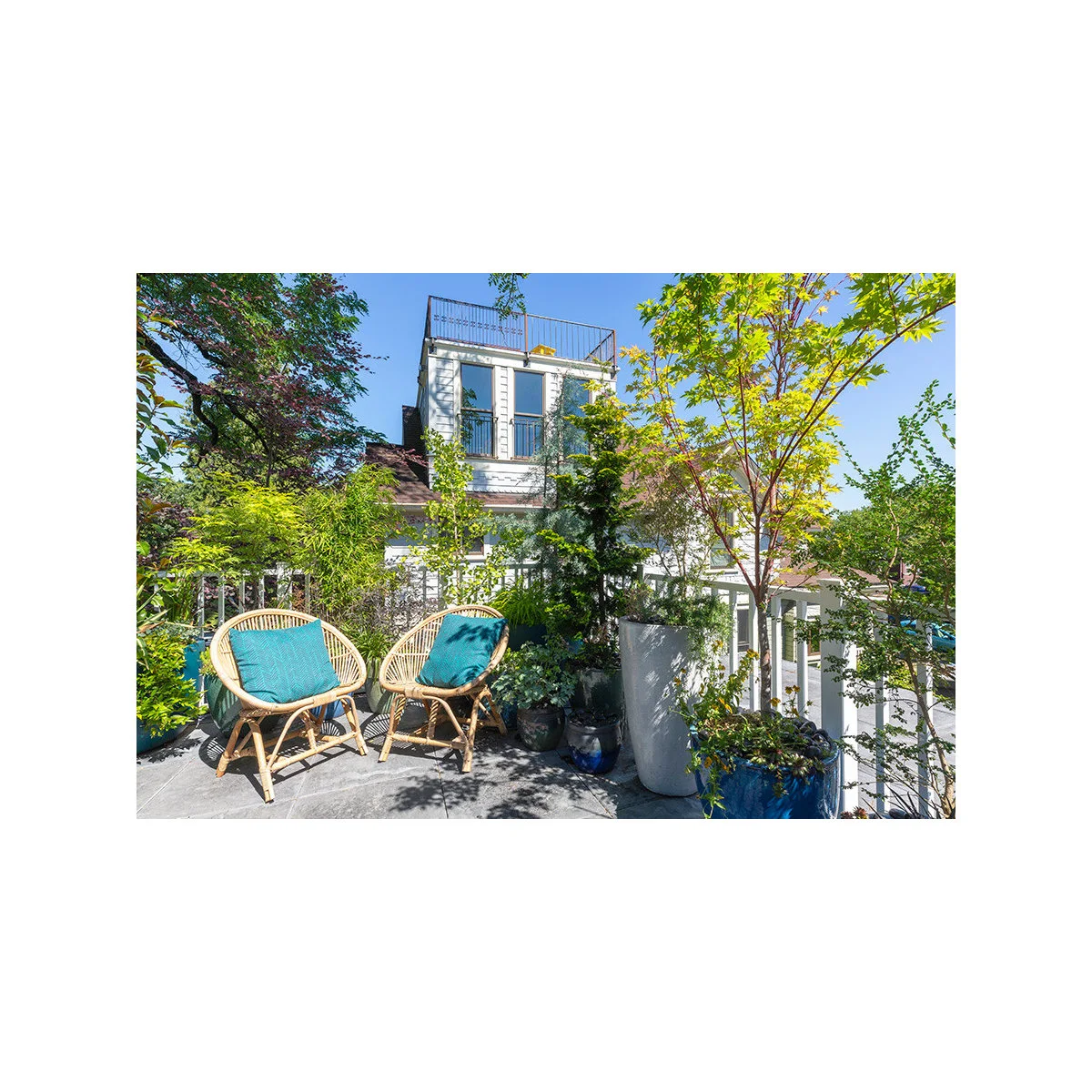 Two round-backed chairs with turquoise cushions sit at the end of a balcony surrounded by tall planters