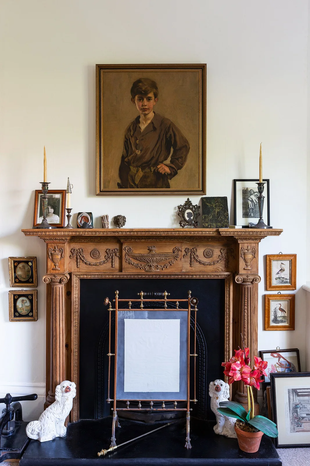 A carved timber fireplace surround stands beneath a portrait with small framed pictures and a decorative screen.
