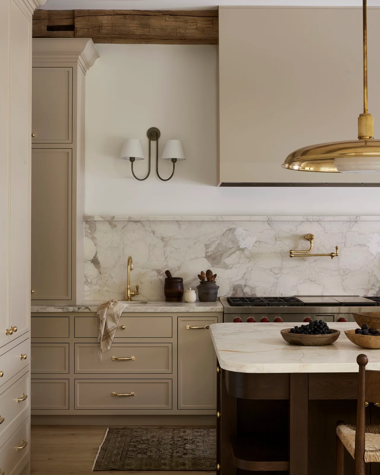 Pale beige cabinets and a matching broad hood surround a veined cooking backsplash beneath an exposed wooden beam
