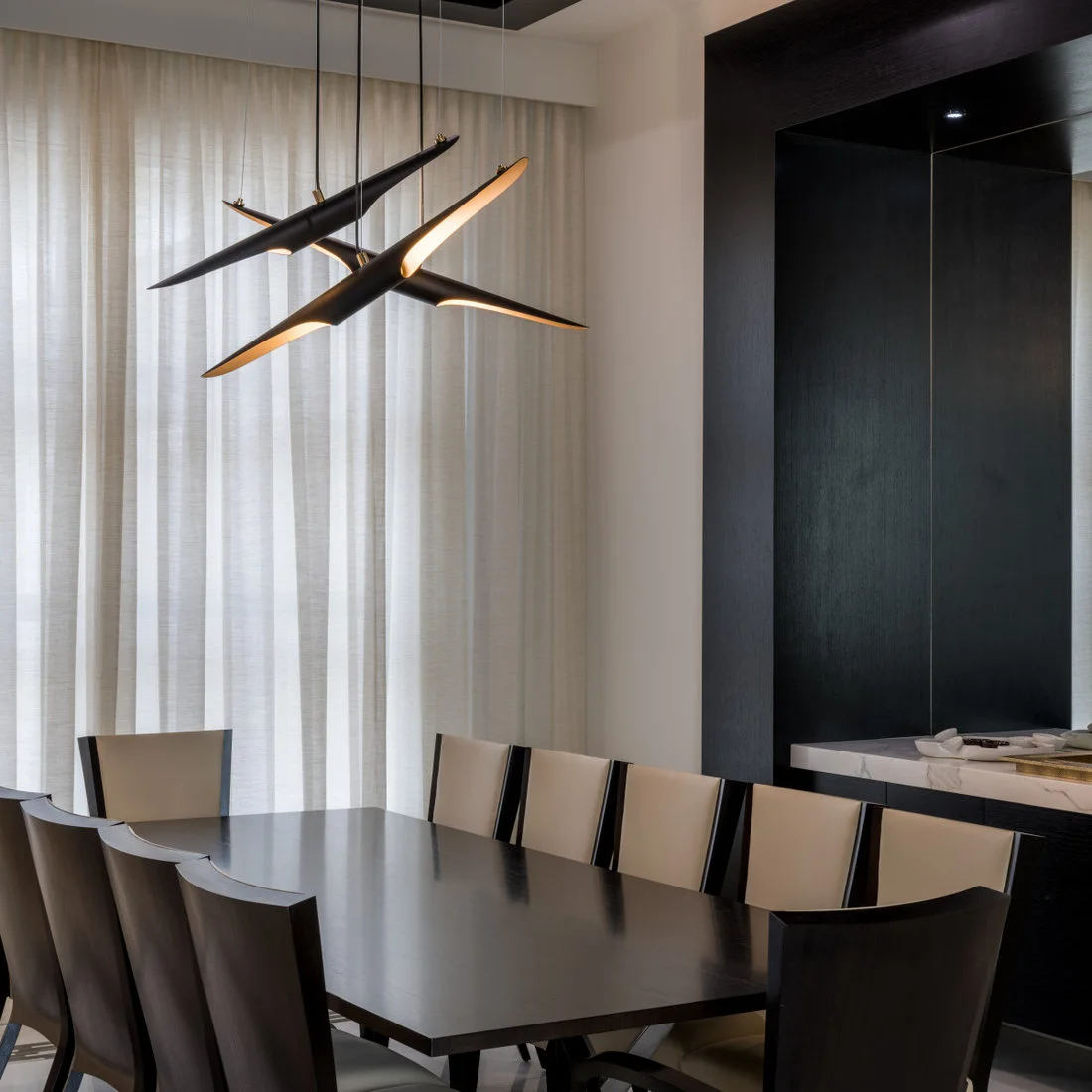 A dark dining table and curved dark chairs with pale back panels stand beneath three angular hanging lights beside pale curtains and dark wall storage