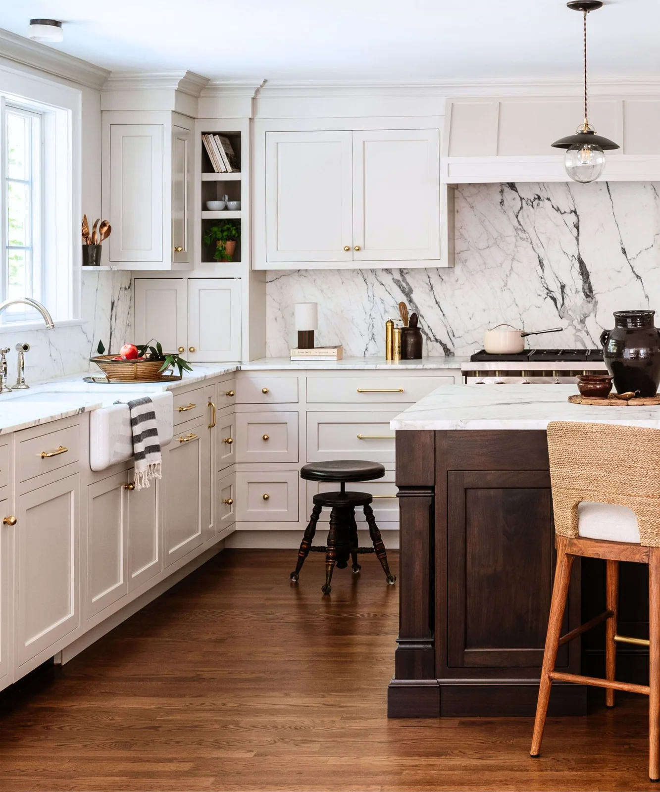 White framed cabinets and a pale veined backsplash meet a dark timber island beside a woven-backed stool.