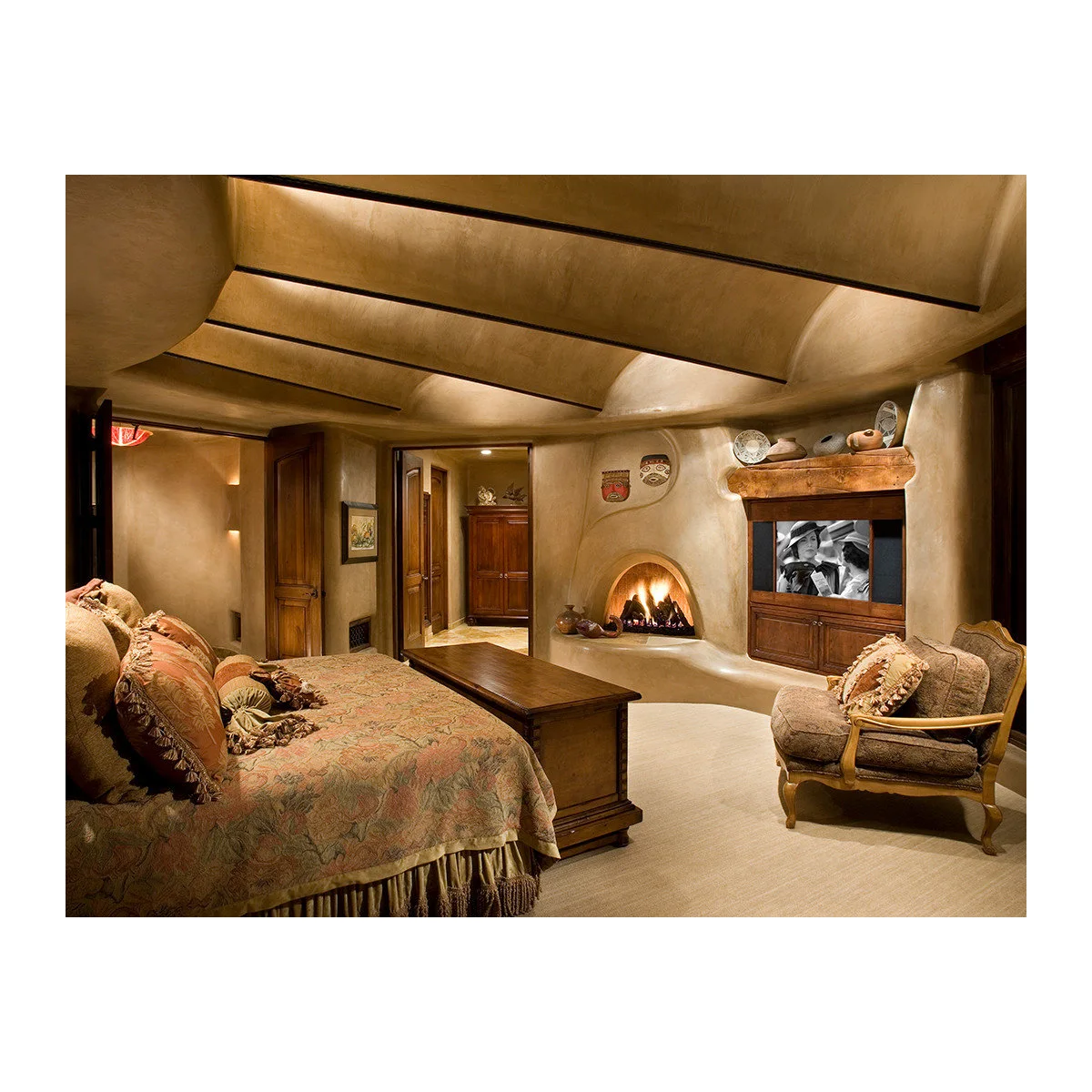 Southwestern bedroom with curved earth-toned walls, an arched fireplace, wood furniture and a small upholstered chair