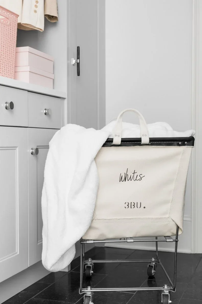 A fabric laundry hamper on a metal wheeled frame stands beside gray cupboards and pink storage baskets.