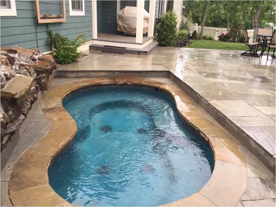A tiny curved blue pool sits within pale paving beside a rock feature and the steps of a covered porch.