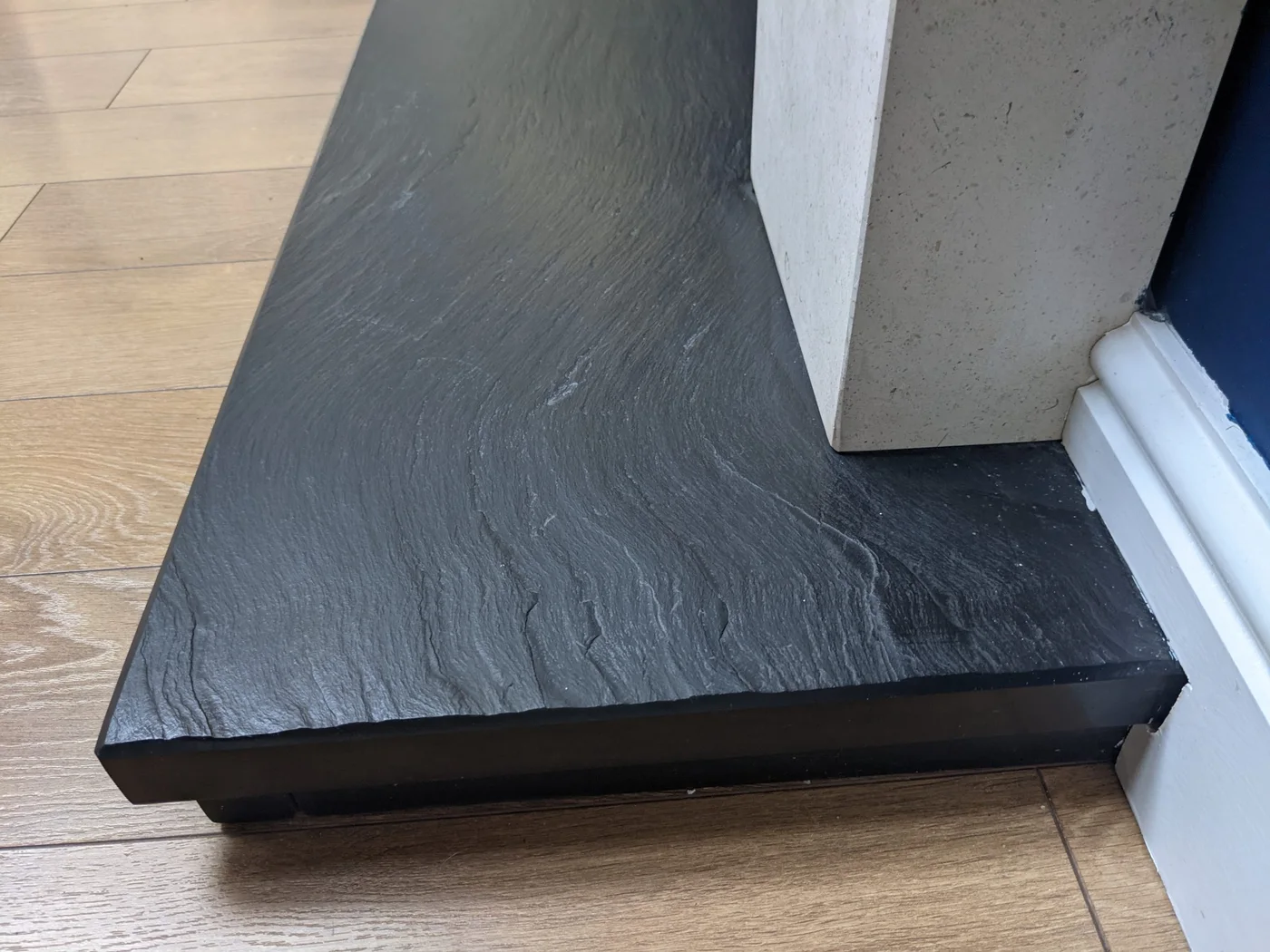 A riven slate hearth meets the upright of a pale fireplace surround above a timber floor.