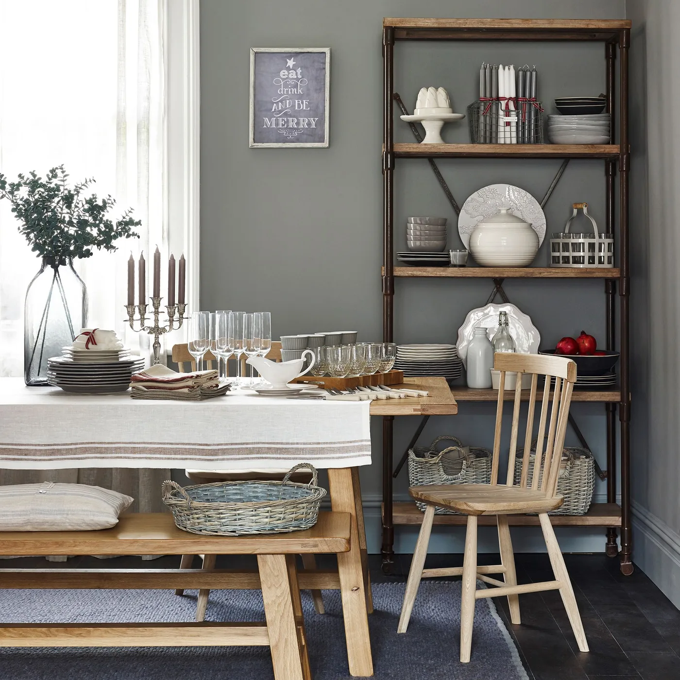 A wood dining table and bench sit beside tall open wood shelves holding pale dishes against a gray-green wall.