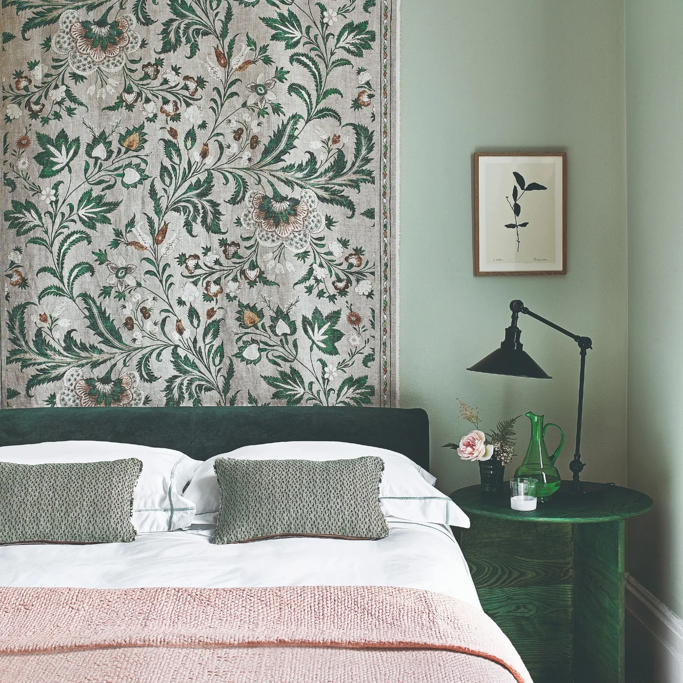 A botanical fabric hanging sits above a dark green headboard beside sage walls, a black reading lamp and white bedding.