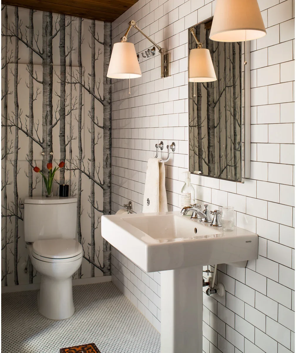 A white rectangular pedestal sink stands against white subway tile, with tree-patterned wallpaper on the adjacent wall and two shaded wall lights.
