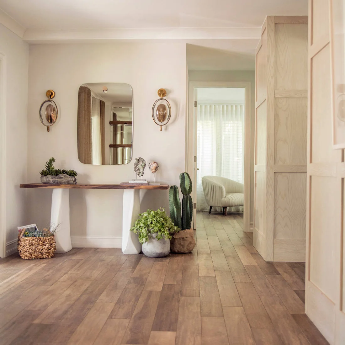 A wood-topped console with pale shaped legs stands below a rounded rectangular mirror between two wall lights beside a wood-paneled doorway.