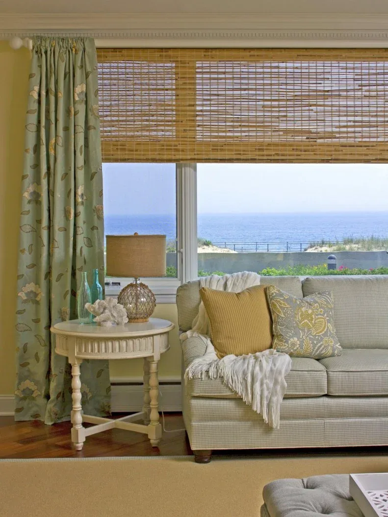 Pale sofa beneath a broad sea-view window with a textured blind, floral curtain and yellow cushions.