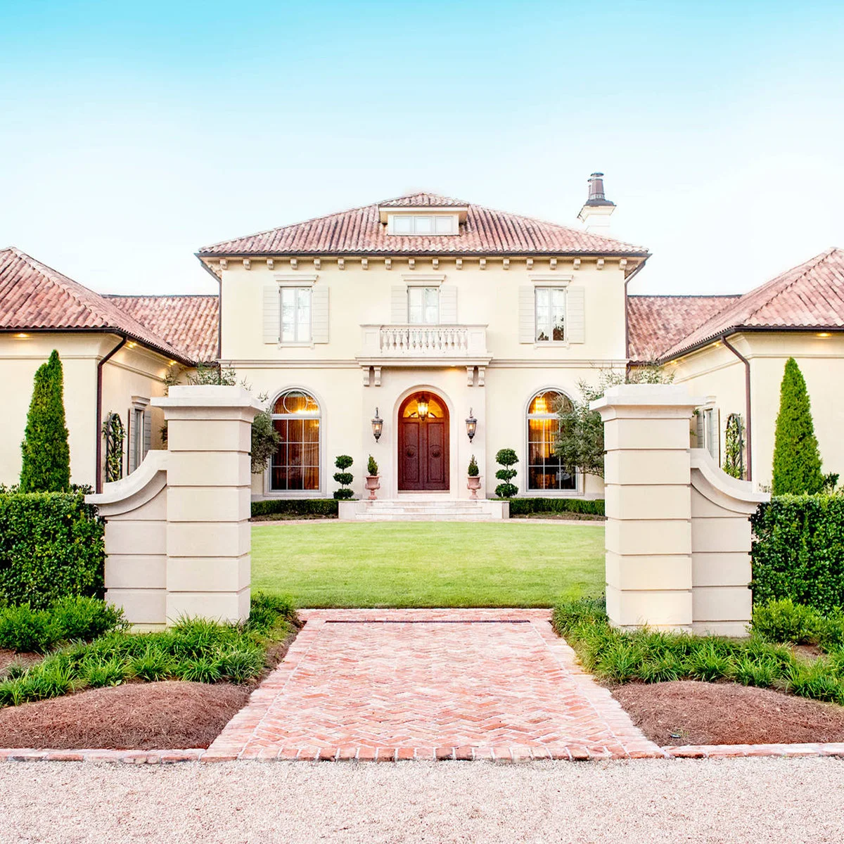 A pale house with an arched front door, narrow clipped foundation hedges and paired sculpted shrubs near the steps.