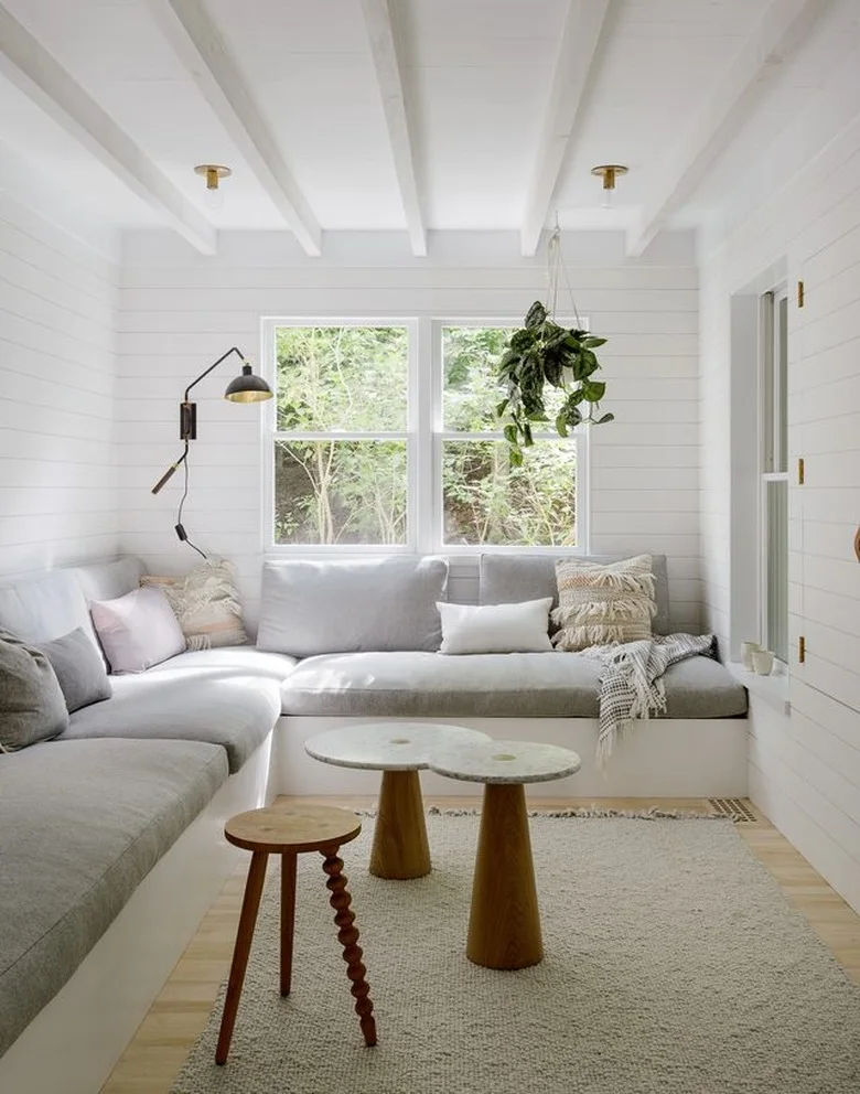 A white paneled living room has low gray corner seating, fringed cushions, a hanging plant and small tables with wood bases.