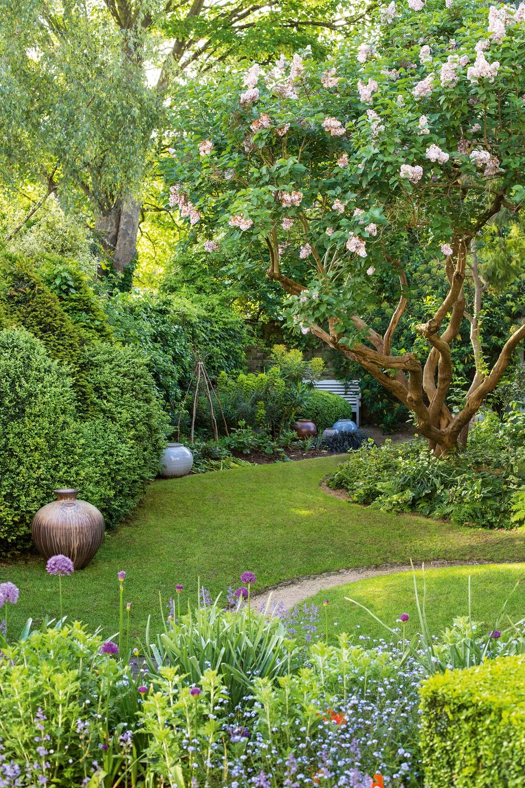 A curved grass route passes a flowering tree and large pots toward a garden bench
