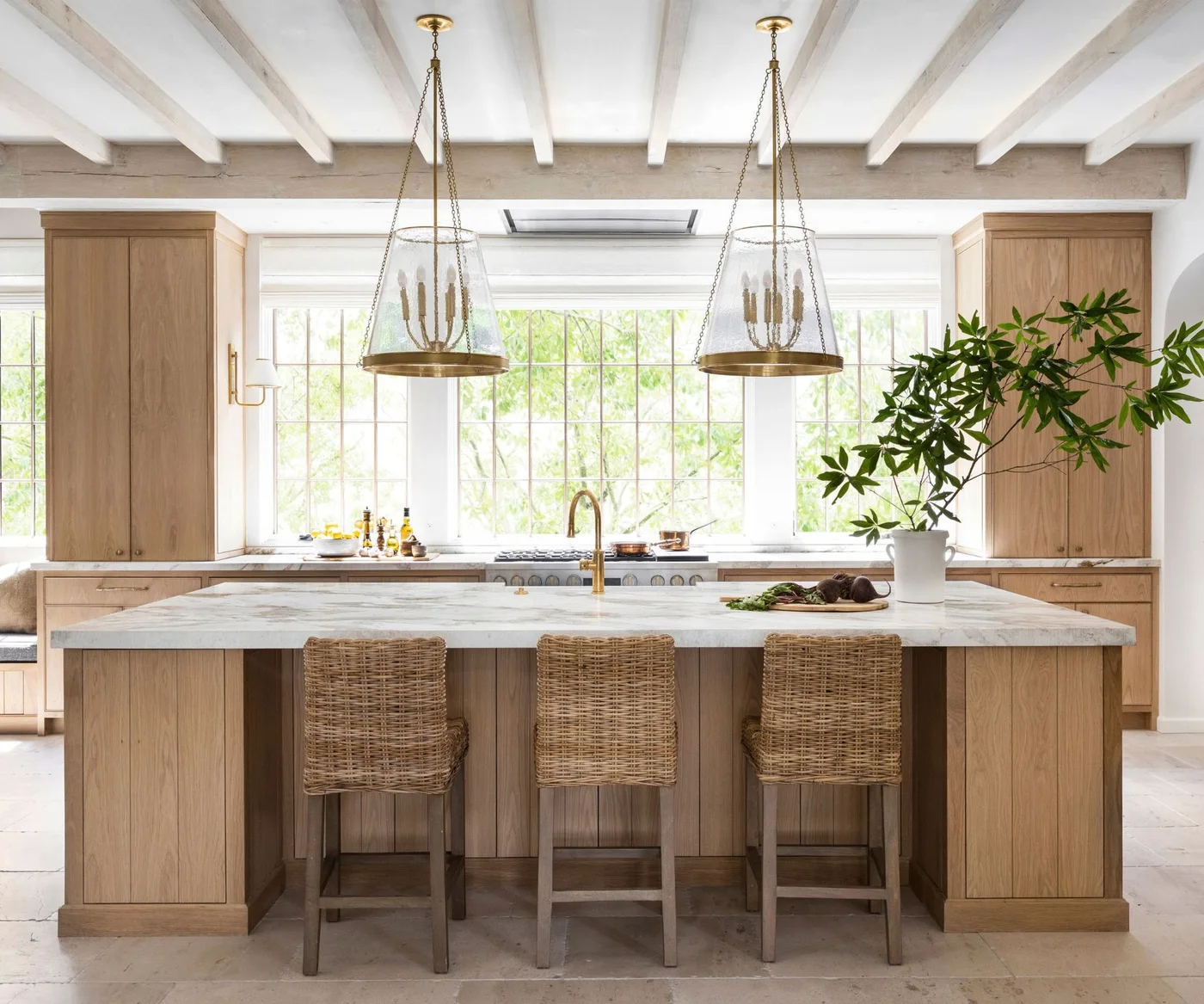 White oak cupboards flank a broad kitchen window behind an island with a pale veined counter and three woven-backed stools.
