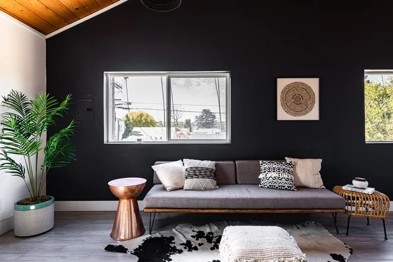 A slim gray sofa with wood trim and narrow dark legs sits against a black wall, with pale cushions, a metal side table and layered floor coverings.