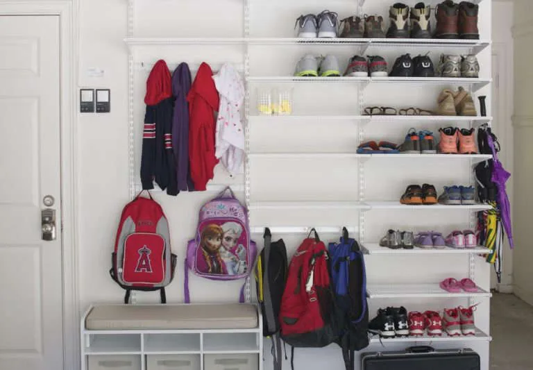 White wall-mounted shoe shelves with coats, backpacks and a small cubby bench beside the entry door.