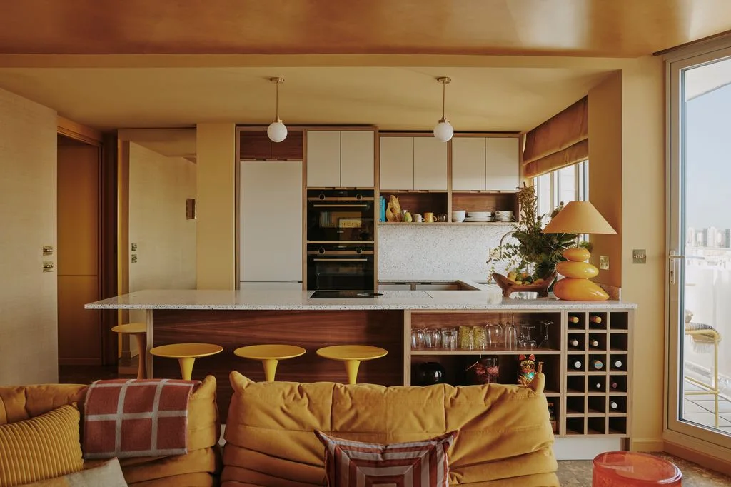 A compact kitchen island has open glassware and bottle storage facing a yellow sofa and matching yellow stools.