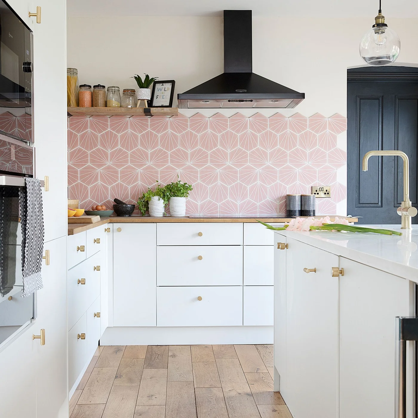 White flat-front cabinets with brass-colored knobs sit beneath pink geometric tiles and a black range hood.
