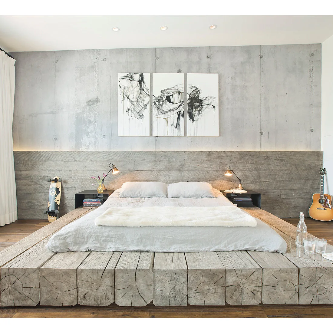 A low bed sits on a broad wooden platform beneath a plank headboard and rough gray wall with black-and-white artwork and small lamps