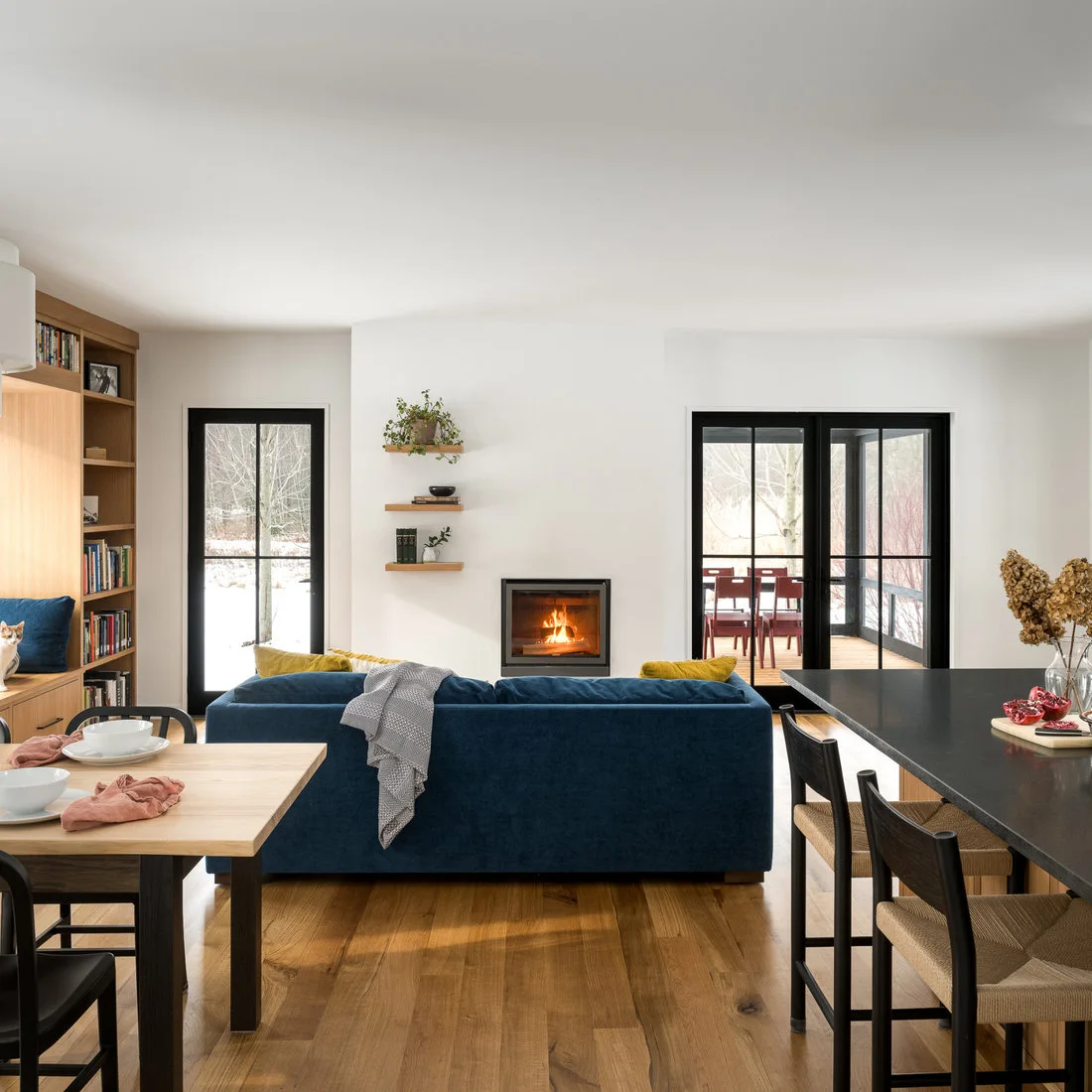 A compact black-framed fireplace sits in a white wall behind a blue sofa, with wood bookshelves and a dining table to one side and a dark counter at the other.