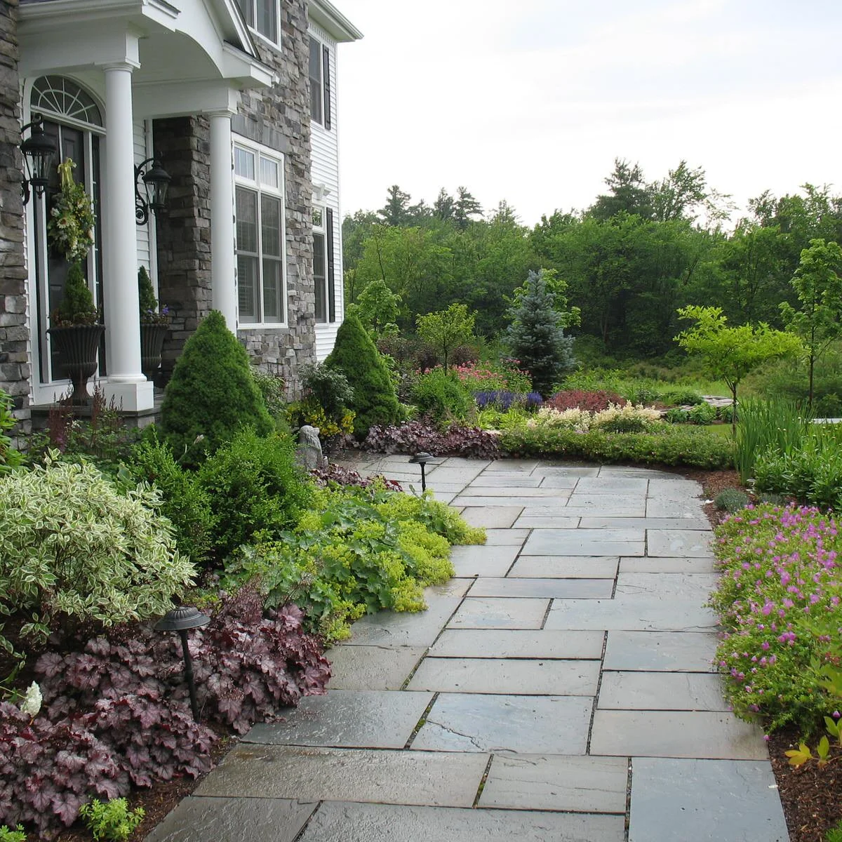 A curved gray path approaches a stone portico beside purple, lime-green and variegated foliage with small pink flowers.