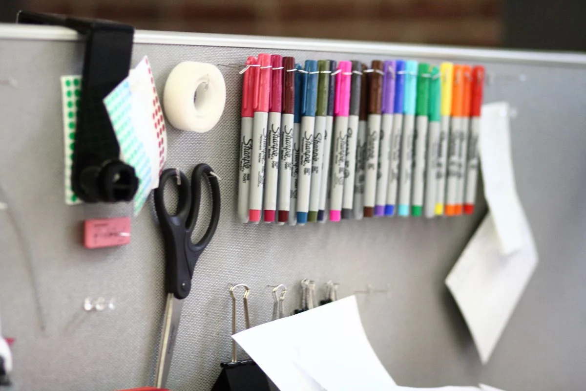 Colored markers, scissors, tape and binder clips held on a gray cubicle panel above the desk.
