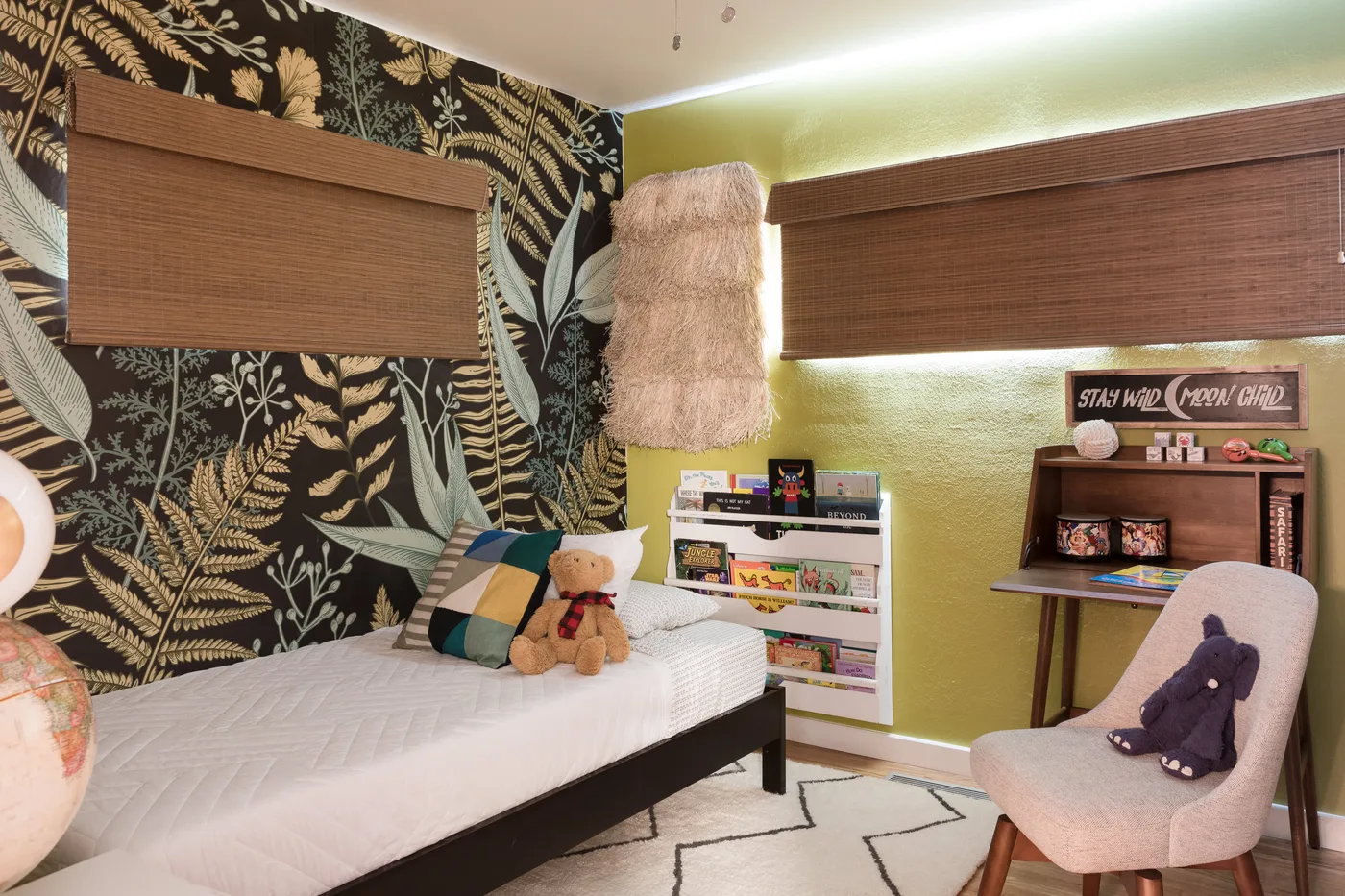 A single white-covered bed stands against dark wallpaper with large leaves and fern shapes beside a vivid green wall, book rail and pink chair