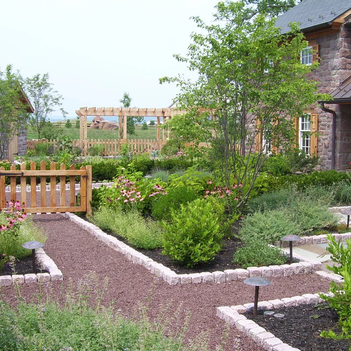 Rough pale edging blocks surround planted beds beside intersecting reddish gravel paths and a wood gate.