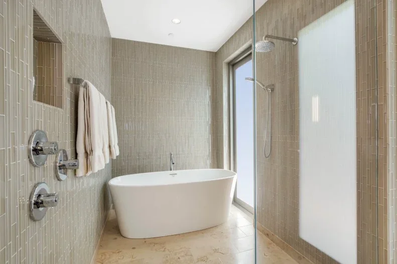 Narrow beige wall tiles surround a white oval tub and shower fittings beside a pale patterned floor and tall frosted window