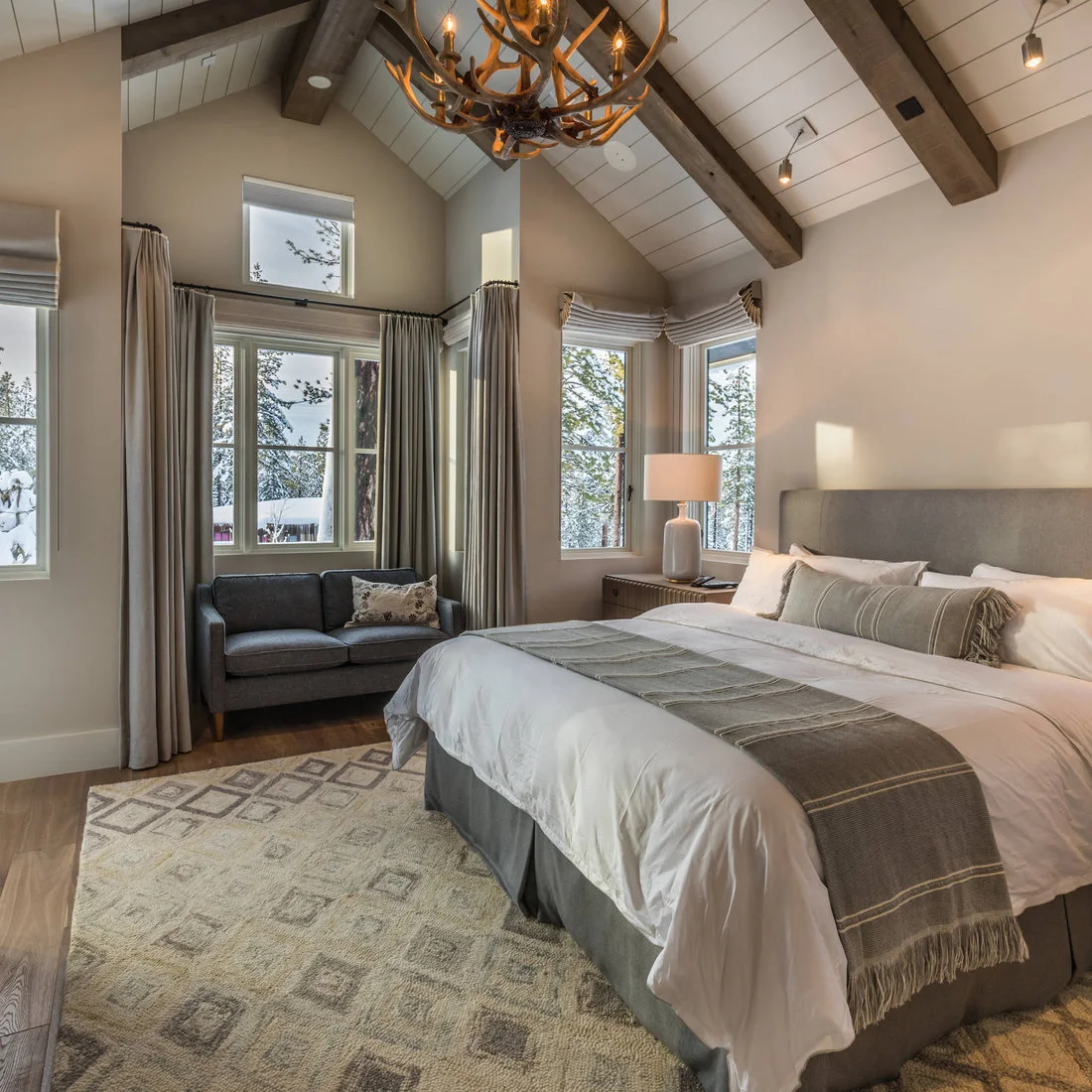 A gray upholstered bed and small sofa by curtained windows beneath white boards and dark beams