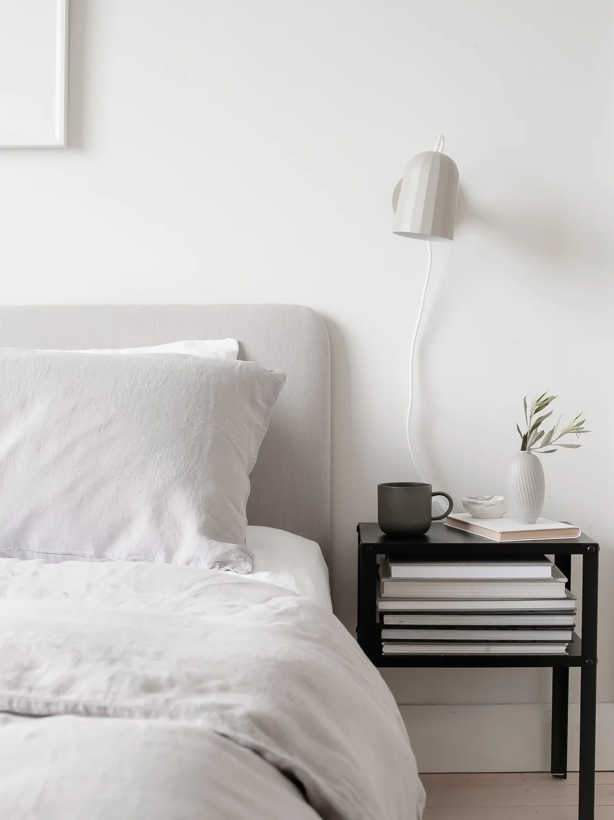 White bedding and a pale upholstered headboard sit beside a small black open table holding books, a dark mug and a white vase beneath a white wall light.