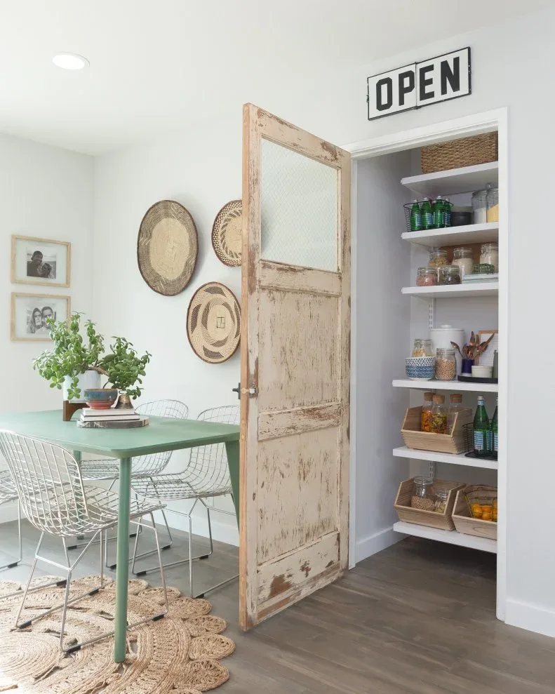 White pantry shelves behind distressed wood glazed door beside green dining table and basket wall display
