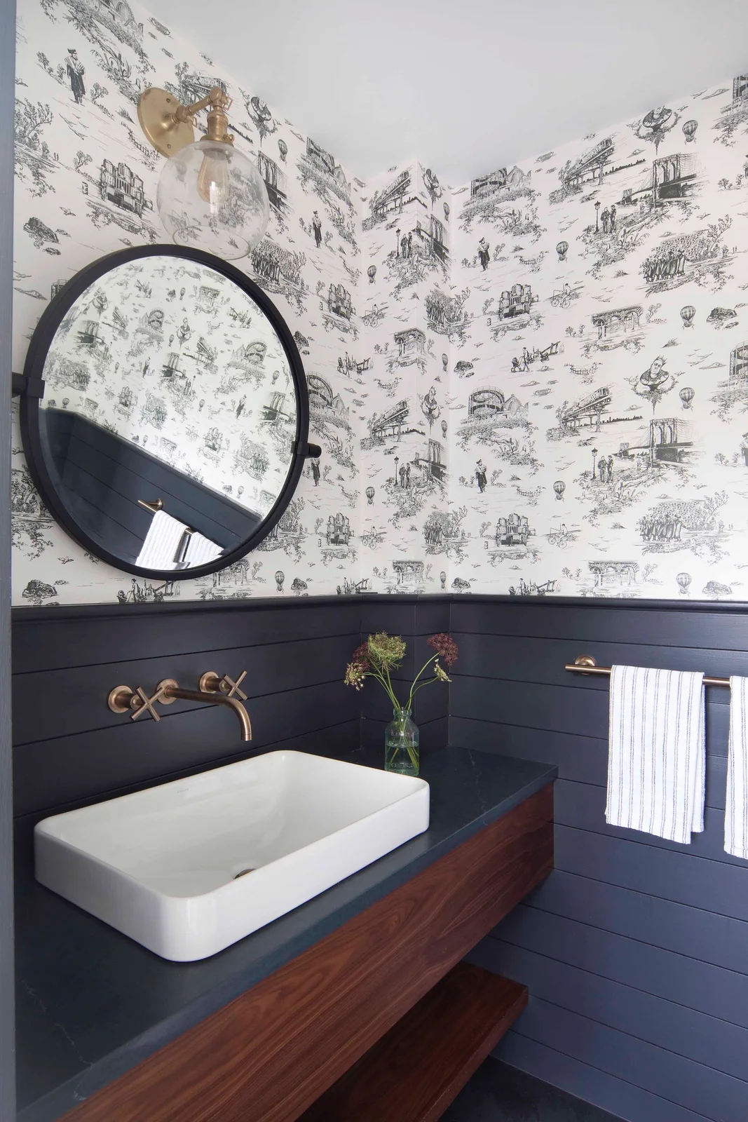 A white rectangular vessel basin sits on a wood vanity beneath a round dark mirror beside navy horizontal boards and black-and-white countryside-patterned wallpaper